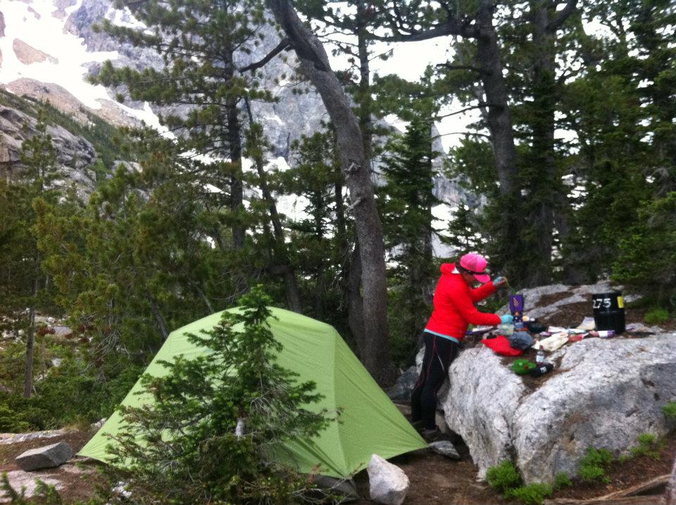 A camper in a red jacket prepares a meal beside a rock and a green tent in Bridger-Teton National Forest campground.