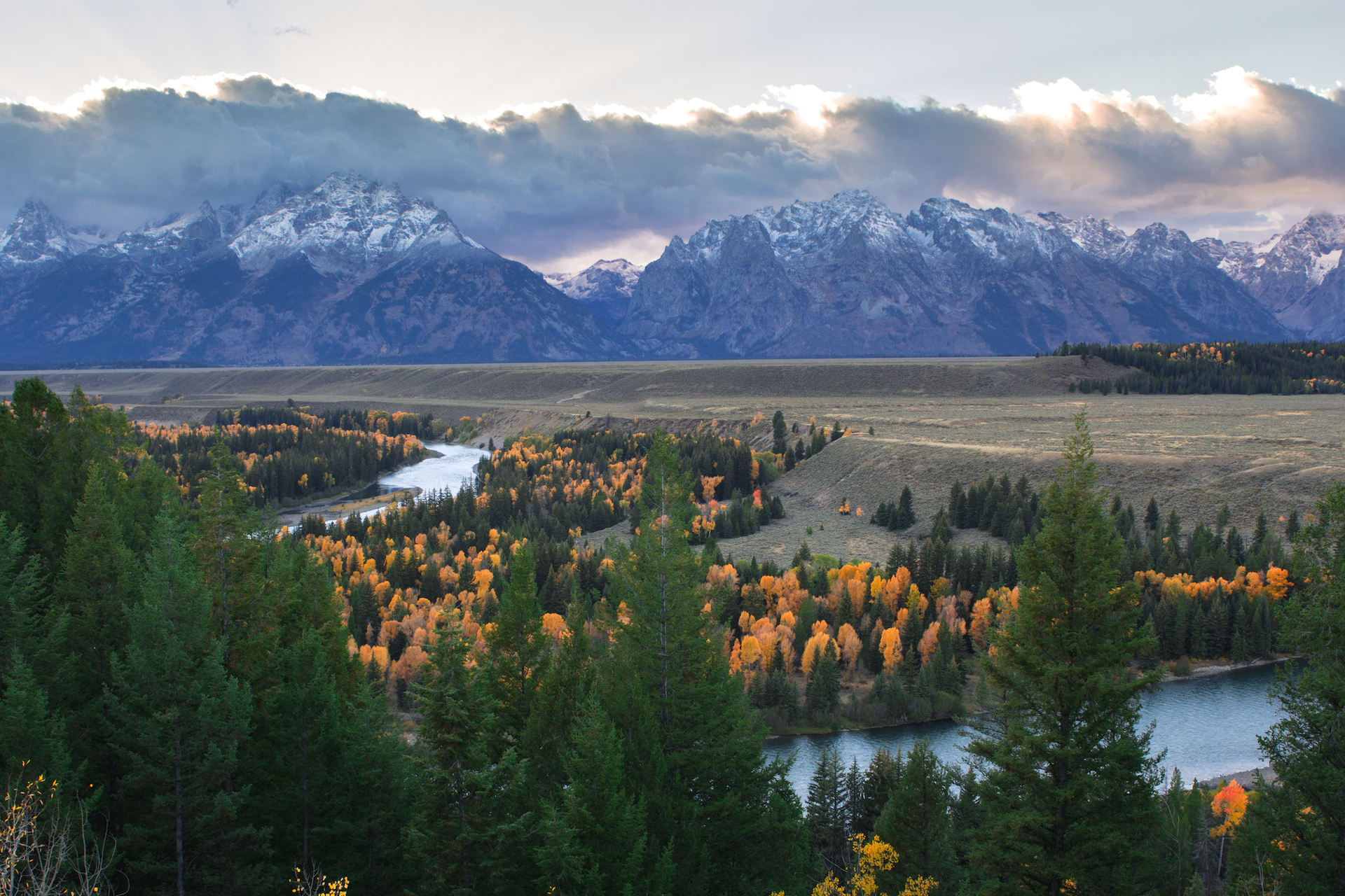 View of Grand Teton mountain range and a valley of trees with autumn foliage along a river
