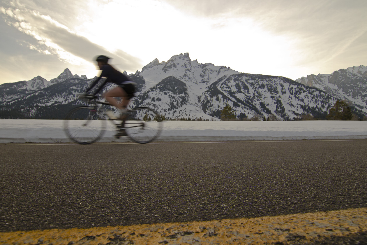 A biker on a road zooms past the snowy mountains of Jackson Hole, WY.