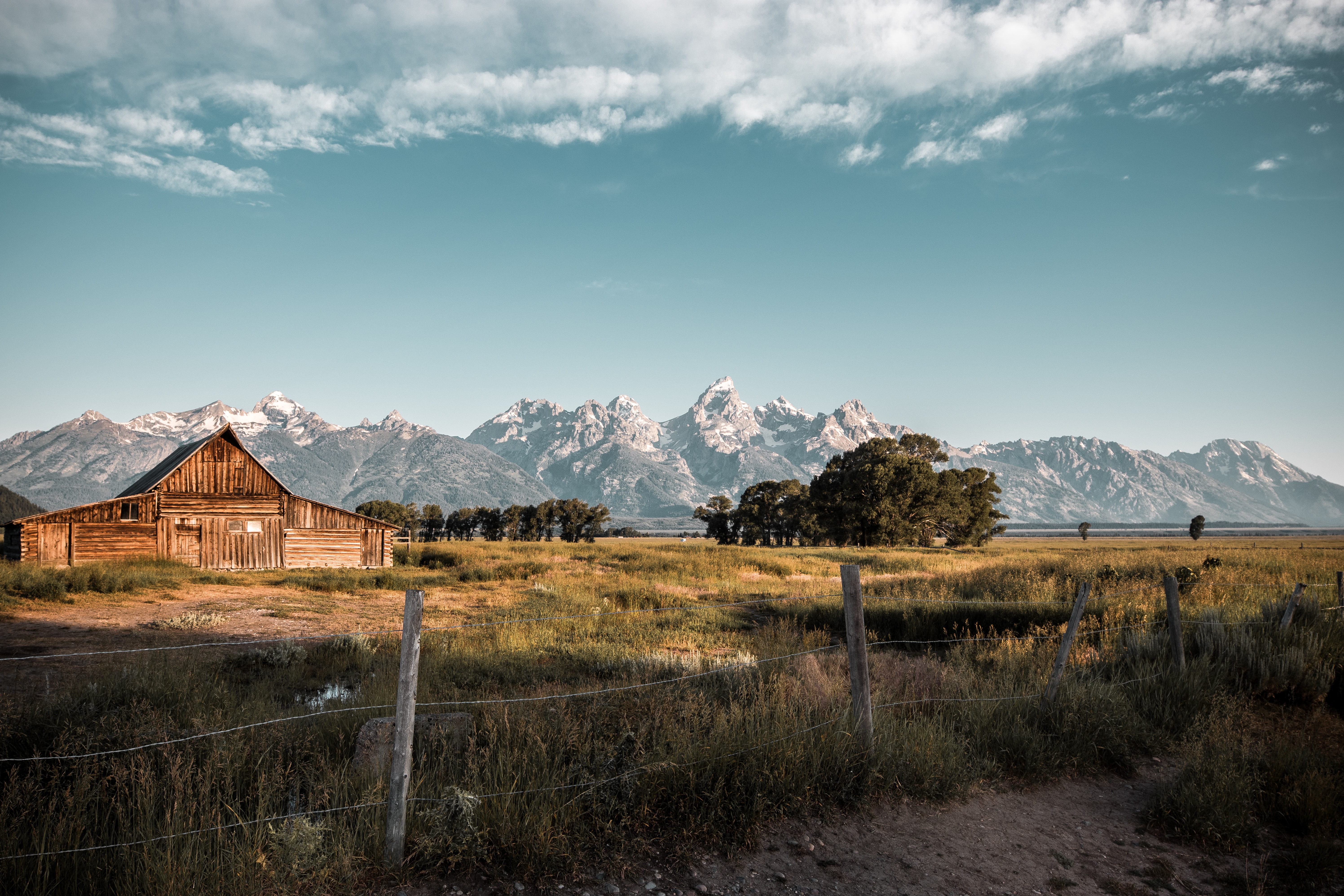 A mildly dilapidated barn with a fence in a field in front of mountains in Grand Teton National Park.
