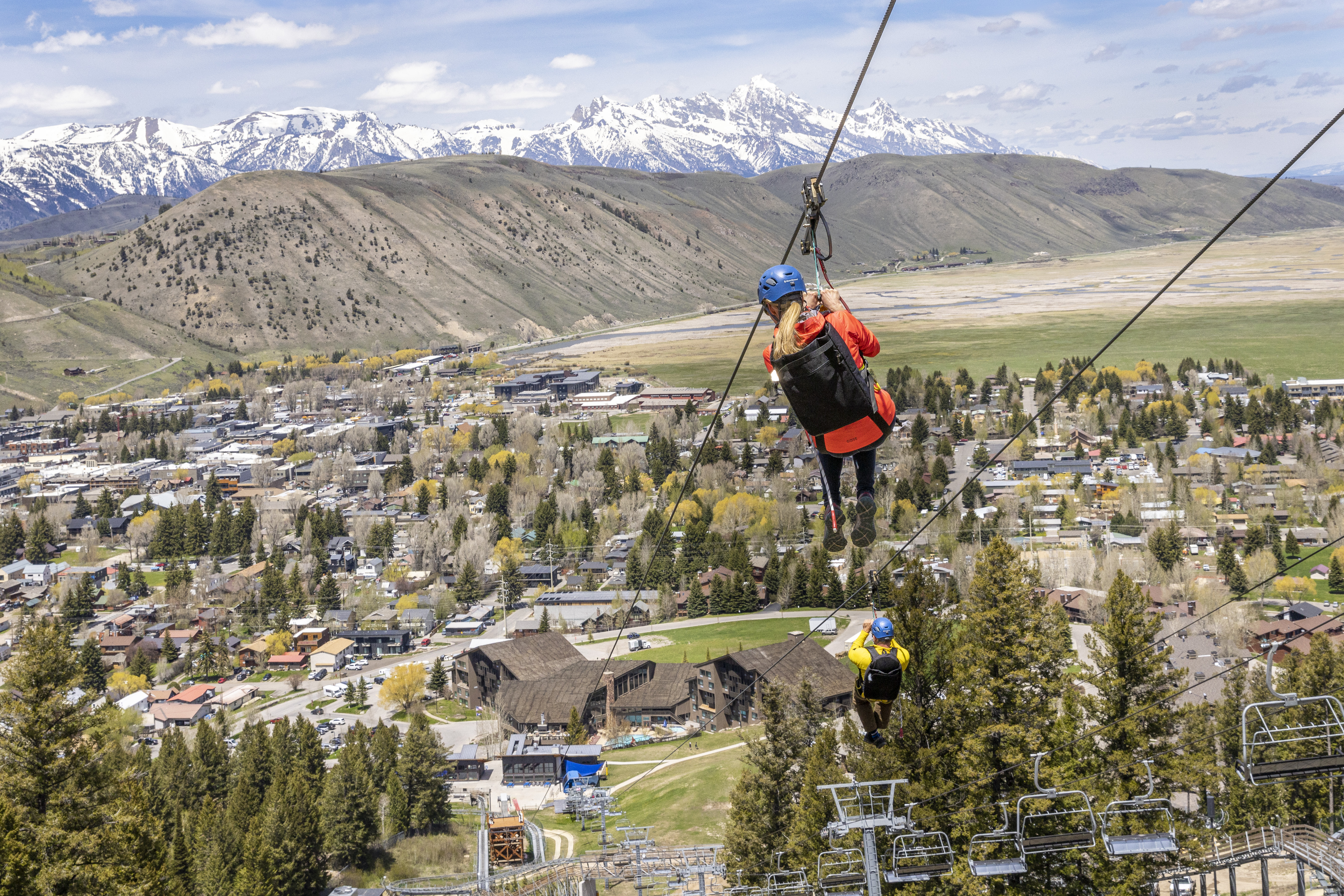 A woman and man descend a mountain on a zipline towards a town with mountains in the distance in Jackson Hole, WY