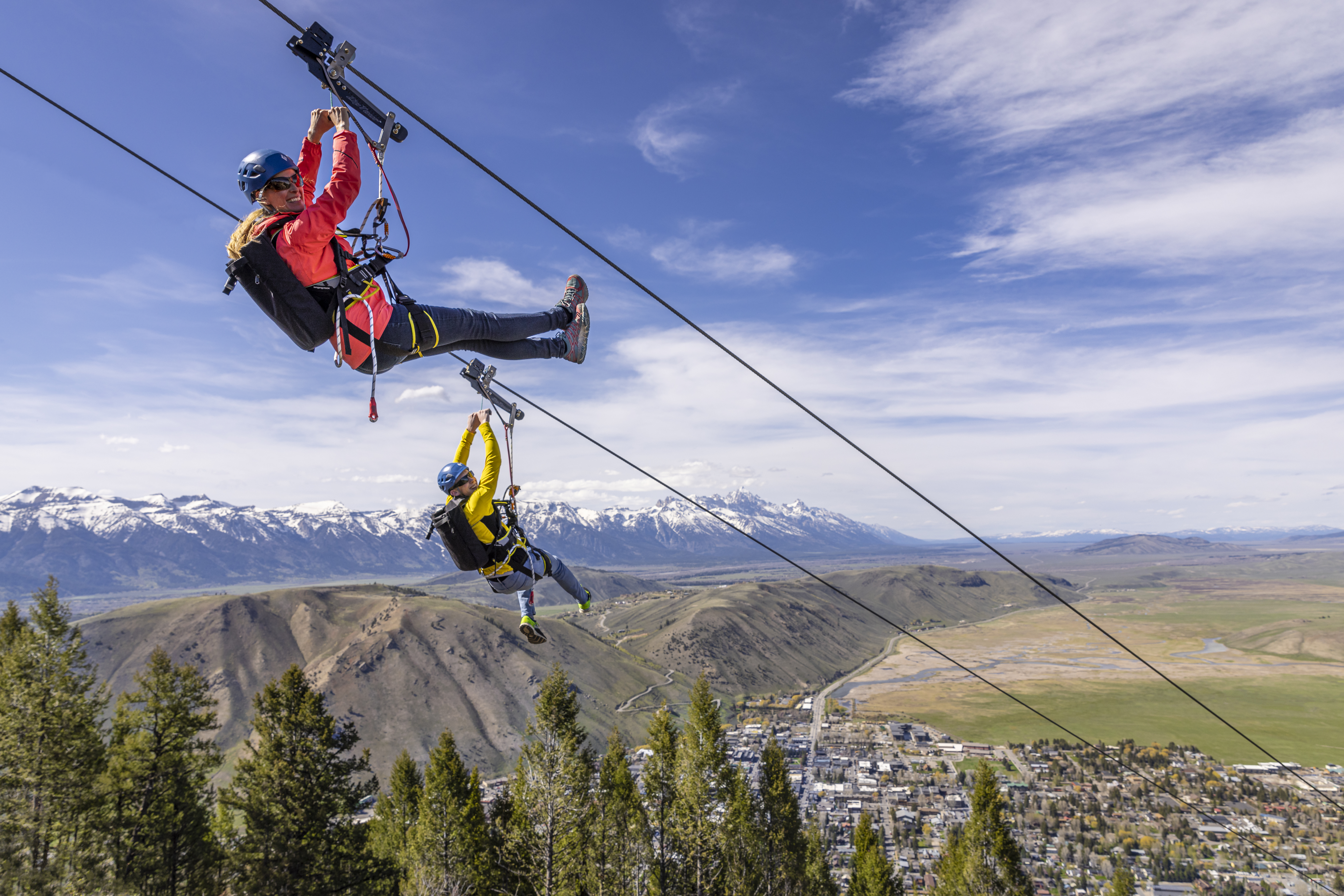 A man and woman glide down a zipline suspended over trees with mountains in the distance in Jackson Hole, WY
